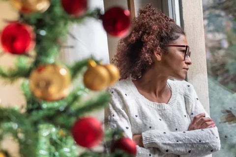 woman with her arms folded looking out a window standing next to a Christmas tree
